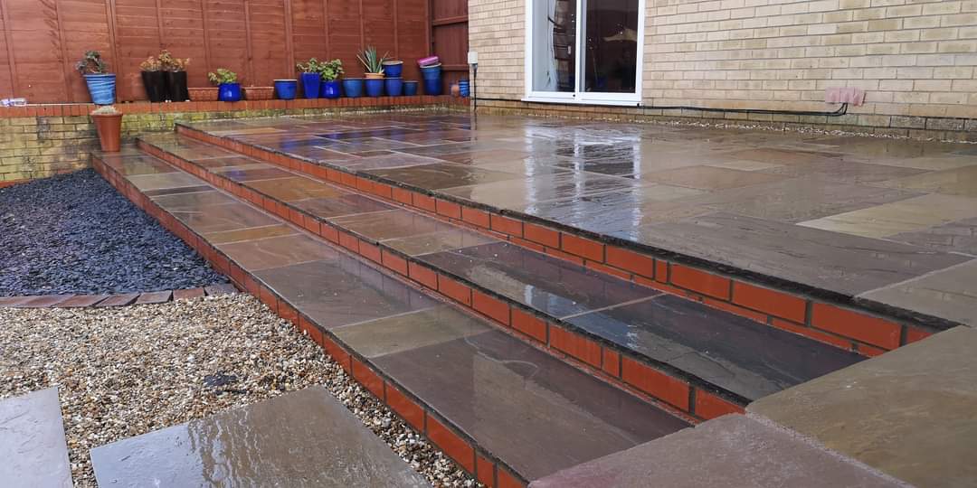 Patio installation Suffolk — Wet paved patio with brick edging and potted plants along fence