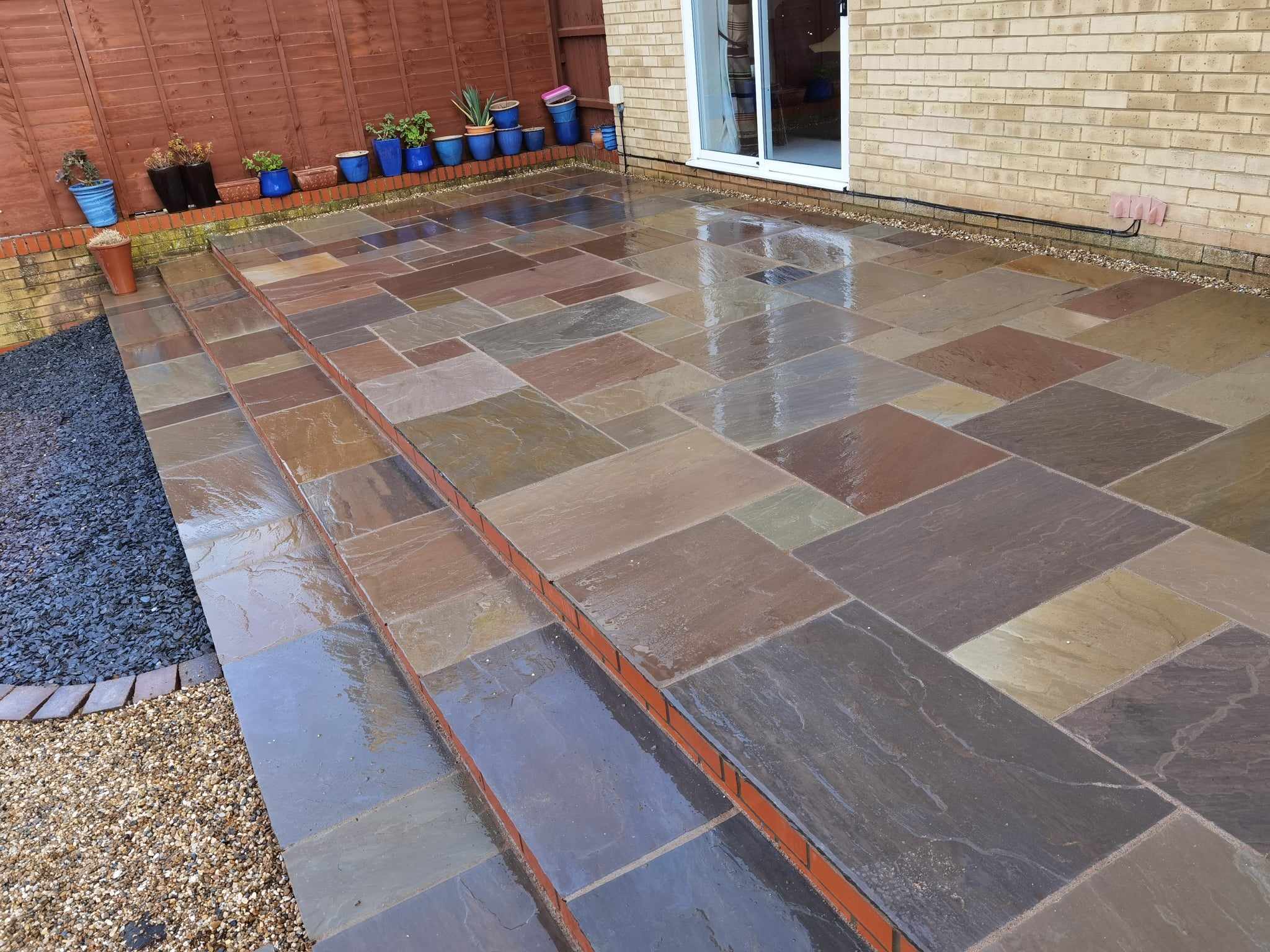 Patio installation Suffolk — Wet slate and stone paved patio area with multiple colored tiles, edged with brick border